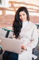 Fashionable female model in elegant white coat sitting at bench holding laptop computer surfing social networks. Professional bussinesswoman sitting at outdoor cafe working with modern computer photo