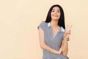 Portrait of cheerful brunette female shows peace gesture, keeps two fingers raised, has pleasant smile, dressed in striped t shirt, poses against studio wall. People and body language concept photo