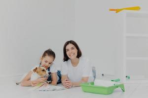 Photo of mother, daughter and pedigree dog lie on floor, take break after painting walls, surrounded with tray and paint roller, isolated over white background, busy with housework and repair