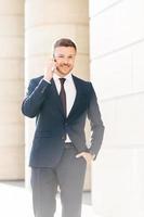 Vertical shot of handsome successful male financial director solves problems via smart phone, looks confidently at camera, keeps hand in pocket, poses outdoor. People and technology concept. photo