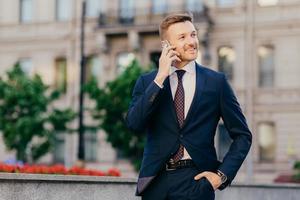 People, conversation and business concept. Handsome prosperous young male entrepreneur makes phone call in roaming, uses tariffs, stands against city blurred background, discusses something. photo