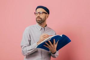 Shot of pensive unshaven man concentrated aside, thinks about what to write. Handsome male architect holds book and crayon, underlines information for project, isolated over pink background. photo