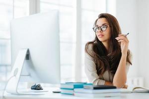 Dark haired lady focused into big monitor, sits at desktop, holds pen and writes notes, wears spectacles for vision correction, poses in coworking space. Femlae manager at workplace, makes research photo