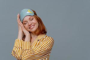 Satisfied ginger woman has nap, sees pleasant dreams, tilts head on palms, smiles gently with closed eyes, wears sleep mask, dressed in striped pajama, isolated over grey background, empty space photo