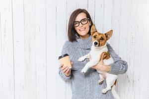 Photo of pleased brunette female carries her favourite dog, drinks hot coffee from paper cup, has pleasant warm smile on face, rejoices nice time spent with pet, poses against white fence background