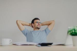 Relaxed handsome male in round glasses, keeps hands behind head, feels relief after making financial project, ready to present information on business meeting, looks thoughfully and gladfully aside photo