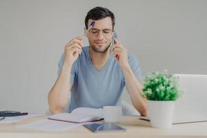 Busy male works on organizational issues, speaks with business partner via cell phone, keeps eyes closed as tries to remember necessary information and write in diary. People and work concept photo