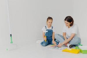 Small girl and her mother sit on floor, look attentively at color palette, choose best colour for painting room, use building tools, pose in spacious white room. Home renovation and design concept photo