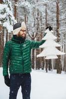 Vertical portrait of dlightful positive bearded male dressed in warm winter clothes, looks with happy expression at artificial fir tree, demonstrates that he protects nature and doesnt cut down trees photo