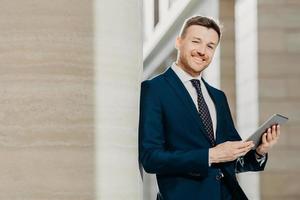 Portrait of stylish male entrepreneur has cheerful expression, holds digital tablet, reads financial news on web page, uses free internet connection, wears formal suit and tie. Career concept photo