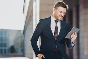 Elegant attractive male executive manager holds modern digital tablet, reads financial news on web page, rejoices having success poses outdoor, has spare time during dinner break. Job and technology photo