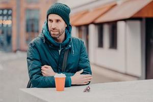 Pensive unshaven young Caucasian man with bristle, wears hat and jacket with hoody, drinks takeaway coffee, stands outdoor, has stroll. People, lifestyle, spare time and street style concept. photo