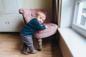 Small blonde child leans at armchair, plays alone at home. Little infant teaches to go. Adorable kid stands near chair, going to play mischievous tricks. Chilren, childhood and lifestyle concept photo