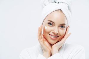 cierra el retrato de una mujer alegre con belleza natural, toca las mejillas suavemente, aplica crema facial para la piel suave, usa una toalla de baño en la cabeza, se viste con una túnica, se para en el interior. tratamiento de belleza, estilo de vida foto