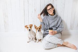 Happy brunette female dressed in loose domestic clothes, being glad, sits on floor, relaxes with hot cappuccino in paper cup, spends time with dogs, isolated over white fence background photo