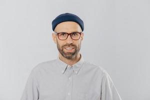 Horizontal view of pleased smiling man wears glasses, hat and shirt, being satisfied with good news, models against white background. Unshaven hipster rejoices new purchase. Fashion concept. photo