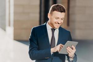 Happy prosperous businessman in formal wear, makes money transaction via touch pad, has glad expression, stands outdoor, connected to wireless intrnet. People, business and technology concept photo