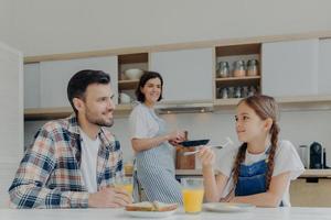 la familia feliz desayuna juntos en la cocina. un niño pequeño positivo habla con su padre mientras come un plato delicioso, la madre se para en el fondo cerca de la estufa, sostiene una sartén, ocupada cocinando. foto