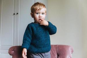 Portrait of handsome blonde little child keeps finger in mouth, looks with his blue appealing eyes, plays alone in his room. Curious small kid stands on armchair, wears warm sweater and trousers photo