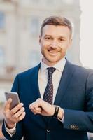 Portrait of well dressed handsome cheerful male executive manager checks time on his wristwatch, uses smart phone, checks email box and waits for bank confirmation, poses outdoor. Vertical shot photo