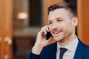 Sideways shot of handsome young male talks in roaming via cell phone, has positive expression, looks thoughtfully aside with copy space for business information. People and conversation concept photo