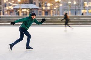 Happy active sporty male involved in winter activities, demonstrates his skating skills on Christmas decorated ice ring, being in movement, has active lifestyle, enjoys his favourite hobby photo