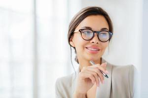 Smiling prosperous lady work in business sphere, holds pen, wears elegant clothes, has healthy skin, minimal makeup, looks somewhere, poses indoor against blurred white background, copy space left photo