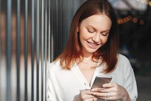 Pleased woman focused into screen of cell phone, checks email box, dressed in white clothes, sends feedback, connected to wireless internet, has brown hair, charming smile, sends text message photo