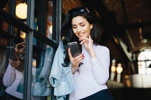 hermosa mujer morena con gafas de sol vestida formalmente usando un teléfono celular coqueteando con un hombre guapo en Internet, esperando su respuesta, mirando la pantalla con placer y gran alegría sentada en un café foto