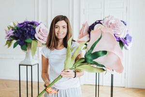 Floral design concept. Beautiful satisfied female florist makes composition of beautiful artificial flowers, decorates hall, has positive smile, dressed casually, being photographed at work. photo