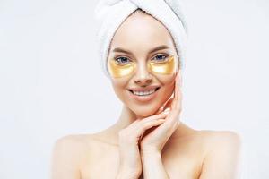 Pretty smiling lady has towel on head touches cheeks, applies golden hydrogel patches, stands shirtless indoor, removes wrinkles, isolated on white wall, has healthy fresh facial skin. High resolution photo