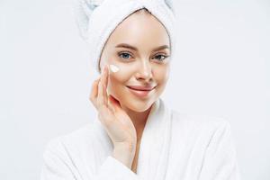 Cropped shot of pretty young woman applies cream for rejuvenation, healthy soft skin, uses cosmetic product, demonstrates nice effect of body lotion, wears comfortable soft white bath robe, towel photo