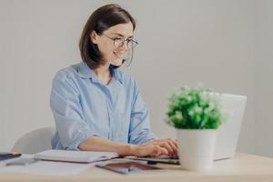 Cheerful successful female freelnacer works remotely, makes research, keyboards on laptop computer, surfes in internet, uses free internet connection. People, business and occupation concept photo