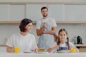 Lovely small kid and mother have breakfast together, sit at kitchen table, eat delicious meal, father stands in background, wears apron and drink coffee. Friendly family gather together at kitchen photo