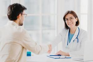 Partnership, assistance, trust and medicine concept. Female doctor shakes hands with thankful patient for good treatment and professionalism, pose in clinic, medical records near on white table photo