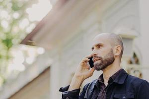 View from above of attractive middle aged male with beard, speaks with business partner via smart phone, poses against blurred background, enjoys using modern technologies. Man has conversation photo