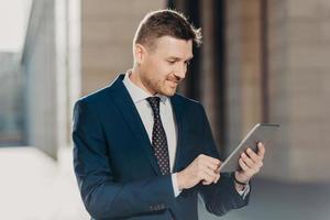 Horizontal shot of successful male office worker dressed in elegant suit, holds modern digital tablet, updates website with financial or business news. Happy trader with modern electronic device photo