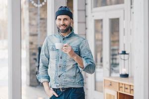 retrato de un atractivo y elegante hombre barbudo con una mirada feliz, usa una chaqueta de mezclilla, sombrero y pantalones de moda, sostiene una taza de bebida caliente, se recrea en casa después de un duro día de trabajo, sonríe agradablemente foto