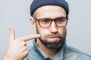 Close up shot of unshaven male with thick beard and mustache blows cheeks and indicates with fore finger, being concentrated on something, isolated over white background. Facial expressions. photo