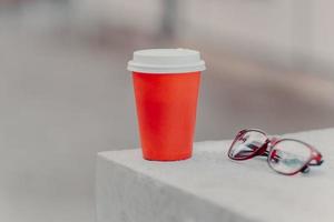 Horizontal shot of red coffee to go and optical glasses. Aromatic coffee for you to drink. Drinking concept. Paper cup of hot beverage photo
