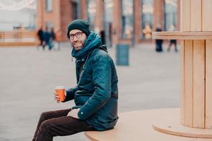 Horizontal view of cheerful European man with thick bristle, wears hat and jacket, holds disposable cup of coffee, breathes fresh air, poses at street. Male model enjoys fresh beverage outside photo