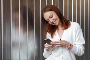 Positive successful European female economist makes money payment on website, poses with cell phone, dressed in white outfit, smiles happily, sends text message, enjoys online communication. photo