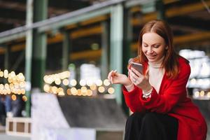Satisfied young European woman in red coat, holds cell phone, reads notification, connected to wireless internet, has spare time, poses outdoor, waits for someone, enjoys online communication