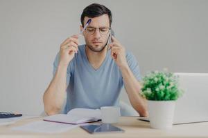 Concentrated young male manager makes internet project, has phone conversation, browses information on laptop computer, tries to thinks what write, isolated over grey background. Job concept photo