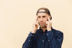 Pensive European male keeps fore fingers on temples, tries to concentrate and recollect his mind, isolated over beige studio background, dressed in fashionable clothes. Thoughtful hipster guy indoor photo