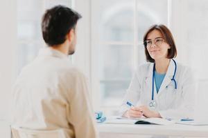 Doctor and patient discuss something, sit at table in clinic. Female cardiologist in eyewear gives medical consultation diagnostic, advice for man how to cure disease, pose in hospital room. photo