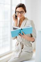 Vertical shot of female administrative manager involved in working process, holds notepad with written notes, discusses something via cellphone, wears spectacles and elegant costume, smiles gently photo