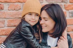 Close up shot of tender pleased young lady smiles gladfully and closes eyes, recieves hug from small daughter who wears leather jackt and hat, pose together over brick wall. Motherhood concept photo