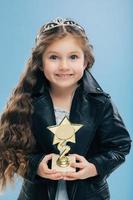 Positive smiling European child with blue eyes, long crisp hair, holds reward in form of star, wears black leather jacket, isolated over blue background. Image of happy girl poses indoor has good mood photo