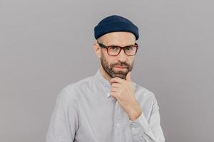 Blue eyed male adult holds chin and looks attentively at camera, ponders on way out or solution, wears spectacles, headgear and formal shirt, models over grey studio wall. Businessman poses indoor photo